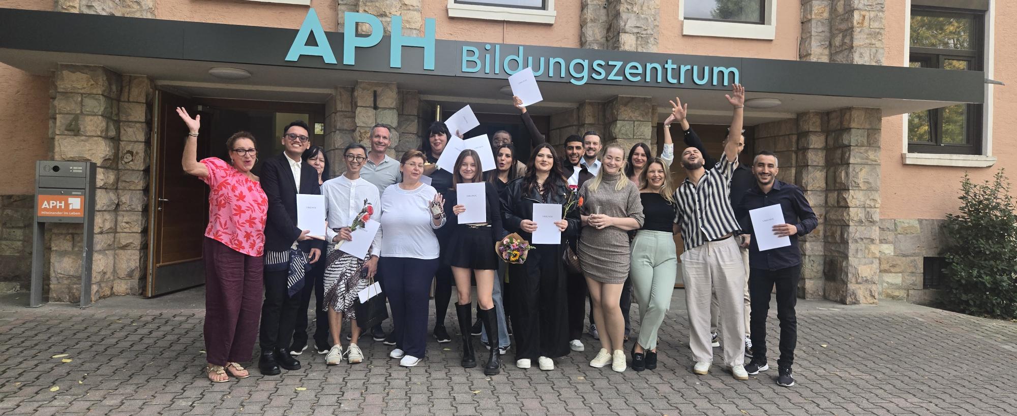 KI-basierte Bildbeschreibung | Eine Gruppe von Menschen steht vor dem APH Bildungszentrum, hält Zertifikate in den Händen und lächelt; einige heben jubelnd die Arme.