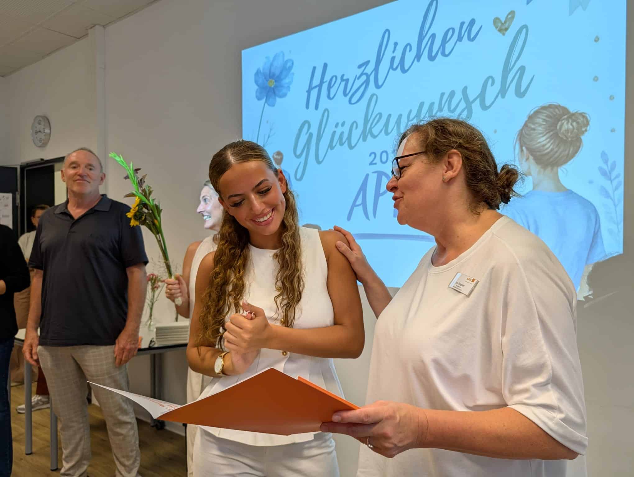 KI-basierte Bildbeschreibung | Eine junge Frau in Weiß erhält vor einem Glückwunschschild mit der Aufschrift „Herzlichen Glückwunsch“ eine Urkunde von einer älteren Frau, während andere zuschauen.