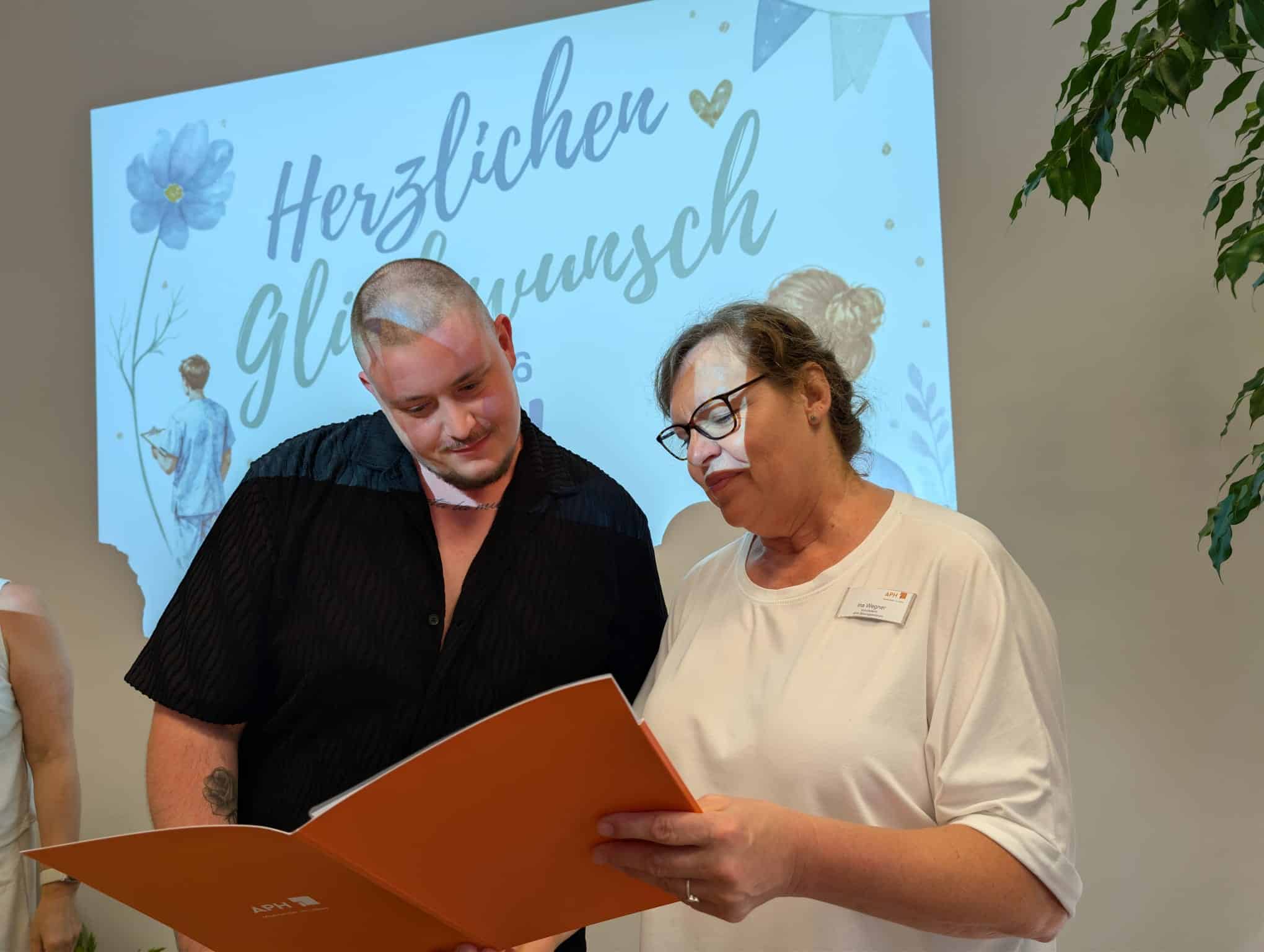 KI-basierte Bildbeschreibung | Zwei Personen stehen nebeneinander und lesen aus einer orangefarbenen Mappe vor, während hinter ihnen eine Glückwunschbotschaft auf Deutsch an die Wand projiziert wird.