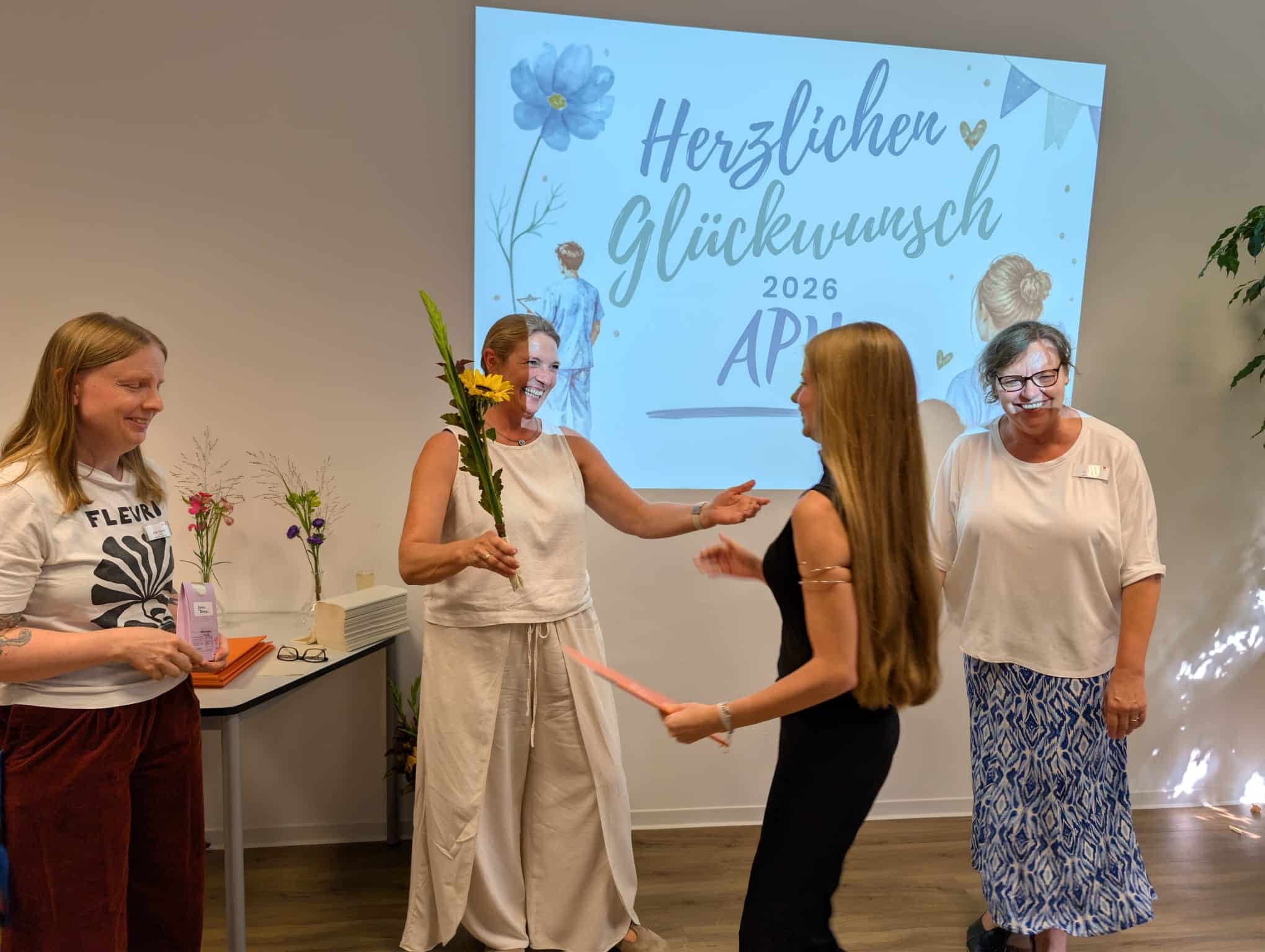 KI-basierte Bildbeschreibung | Vier Frauen stehen gemeinsam in einem Raum, wobei eine einer anderen eine Sonnenblume und ein Blatt Papier reicht. An der Wand hinter ihnen ist eine Glückwunschbotschaft zu sehen, die mit Blumen und Herzen verziert ist.