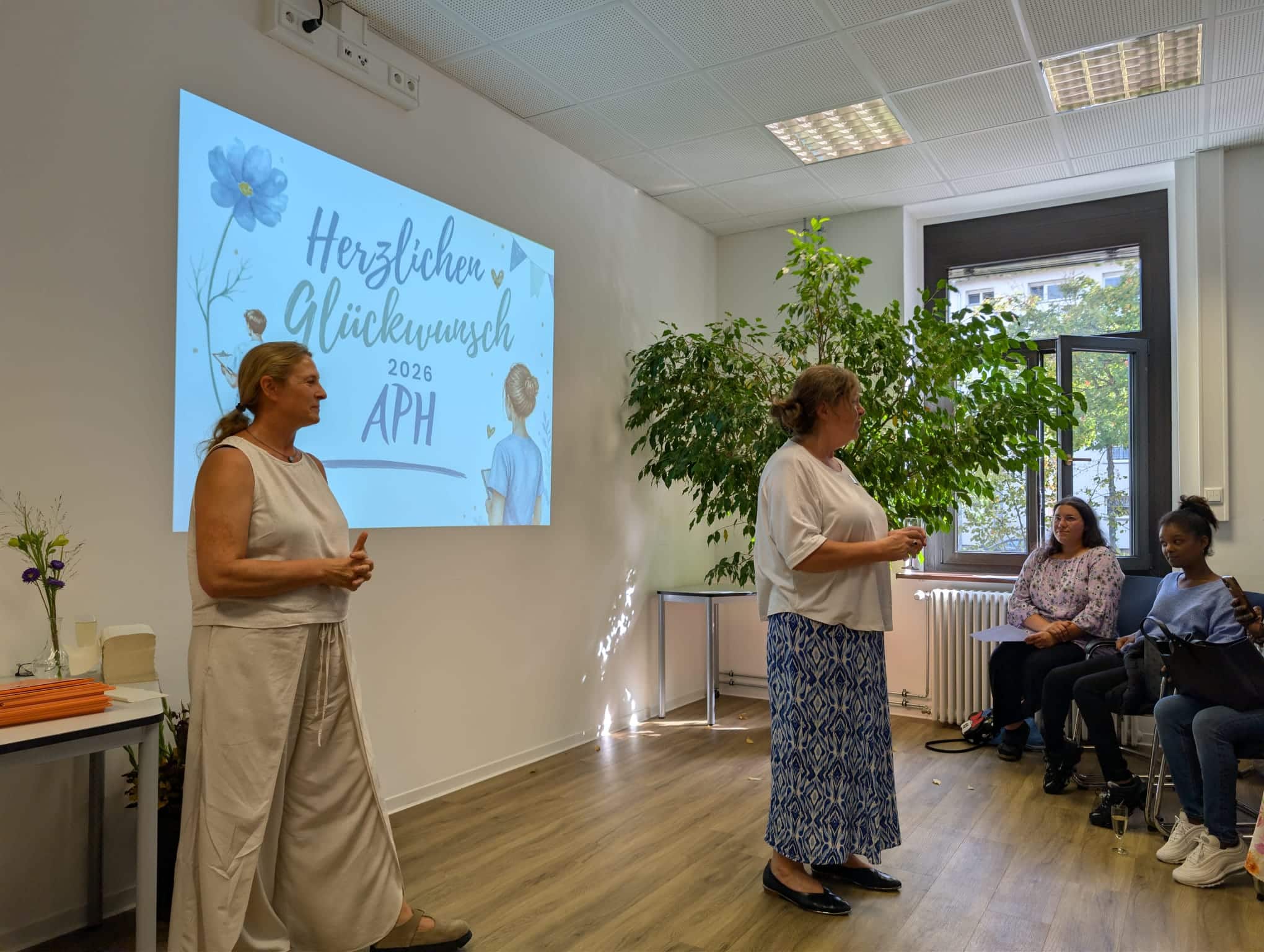 KI-basierte Bildbeschreibung | Zwei Frauen stehen in einem Klassenzimmer und sprechen, während an der Wand „Herzlichen Glückwunsch 2026 APH“ projiziert wird. Am Fenster sitzen mehrere Personen und hören zu.