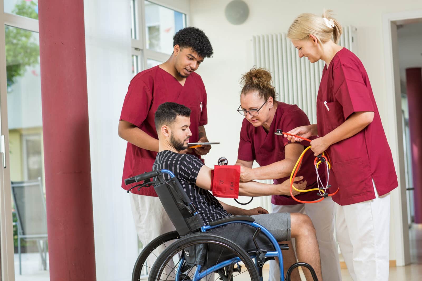 KI-basierte Bildbeschreibung | Drei Angestellte des Gesundheitswesens in roten Uniformen helfen einem Mann im Rollstuhl und messen seinen Blutdruck mit medizinischen Geräten in einer hellen Innenumgebung.
