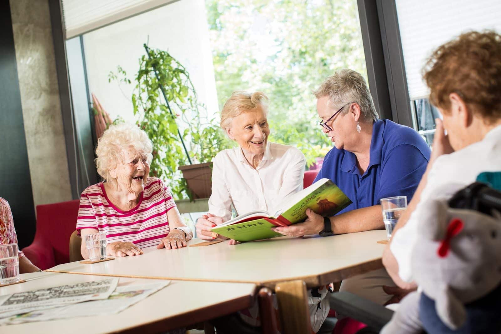 Frauen sitzen um einen Tisch, lachen und eine Pflegerin zeigt etwas in einem Buch