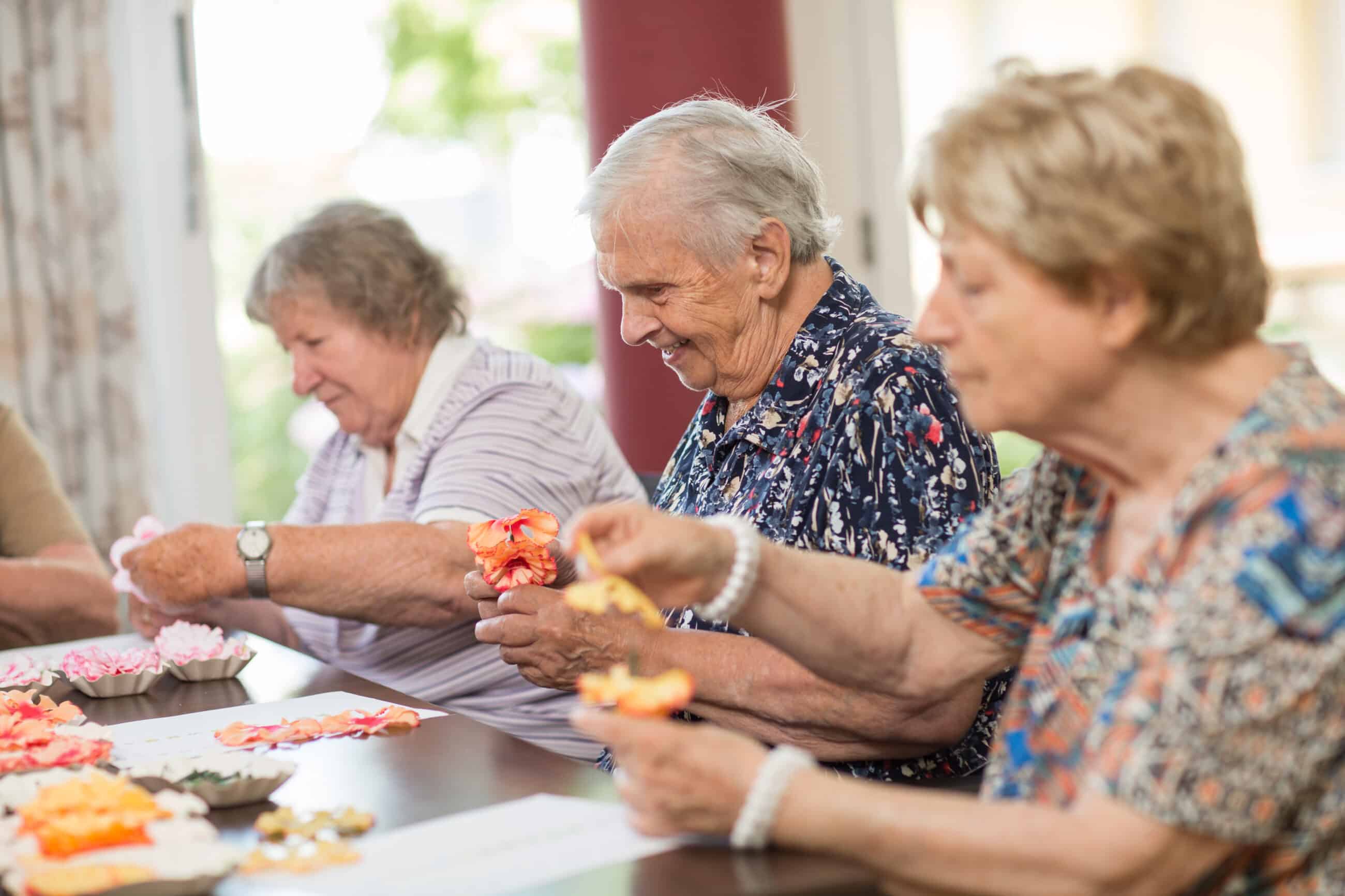 KI-basierte Bildbeschreibung | Drei ältere Frauen sitzen an einem Tisch und basteln mit buntem Papier in einem hellen Raum.