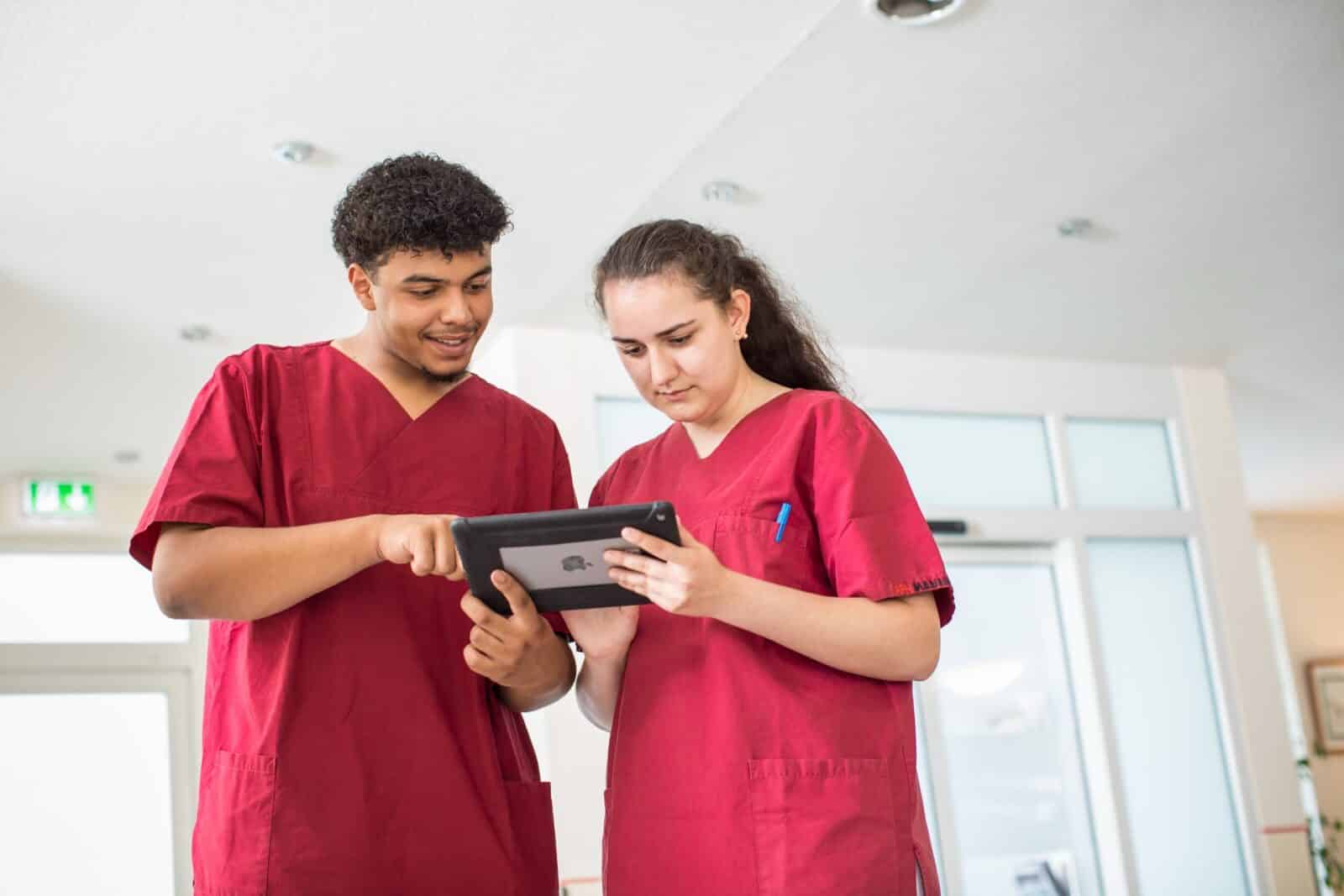 Zwei Auszubildende in roter Pflegekleidung schauen gemeinsam auf ein Tablet