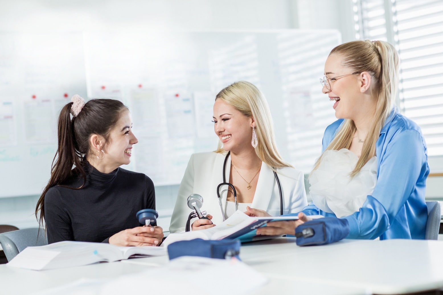 Drei Frauen sprechen lachend an einem Tisch mit Lehrbüchern und Blutdruckmessgerät