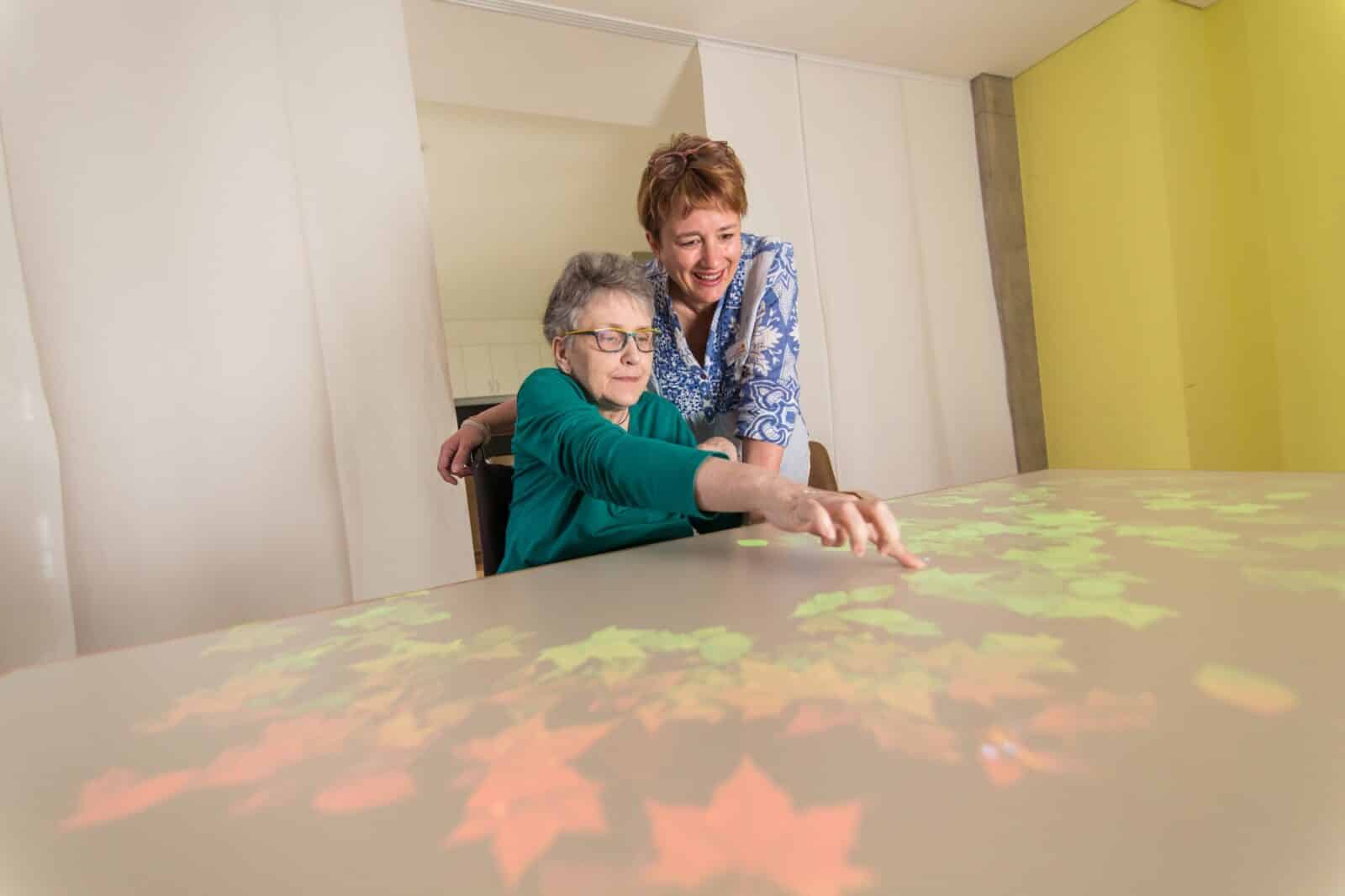 Eine Pflegerin und eine ältere Frau an einem interaktiven Tisch mit projizierten Herbstblättern